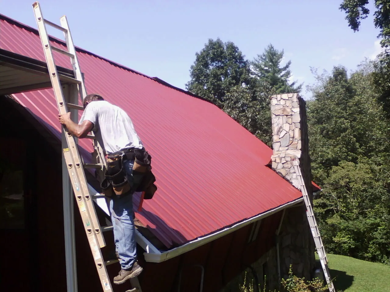 Roofer on a ladder beside a red metal roof and stone chimney — Soffit & Fascia Repair by Roofing Brothers in Chantilly